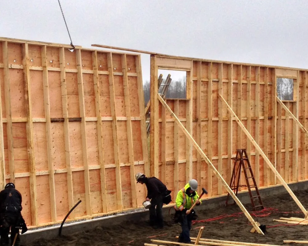Two workers frame the wooden structure of a residential building under construction in a rural area.